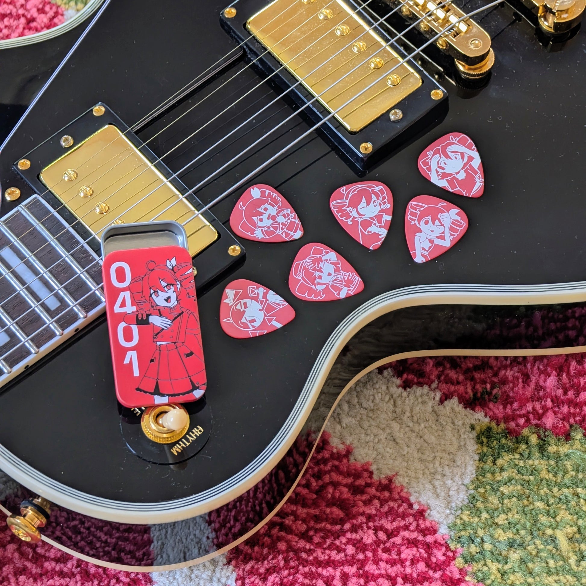 A close-up of a black electric guitar body with gold hardware, with several red guitar picks featuring illustrations of Kasane Teto as she appears in different songs and a red metal tin illustrated with a 3/4 Kasane Teto and the numbers "0401".