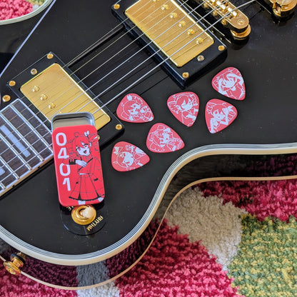 A close-up of a black electric guitar body with gold hardware, with several red guitar picks featuring illustrations of Kasane Teto as she appears in different songs and a red metal tin illustrated with a 3/4 Kasane Teto and the numbers "0401".