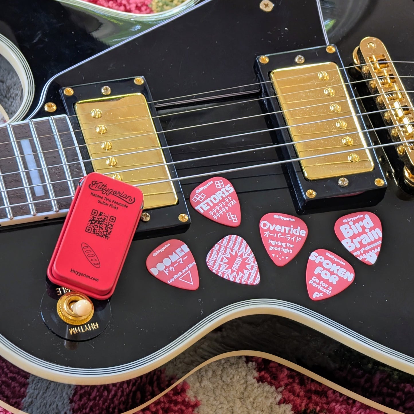 A close-up of a black electric guitar body with gold hardware, with red guitar picks (rear side of previous image) featuring illustrations of songs with Kasane Teto on vocals and the rear side of the red metal tin showing the Kittygorian logo, website, link QR code, and a baguette.