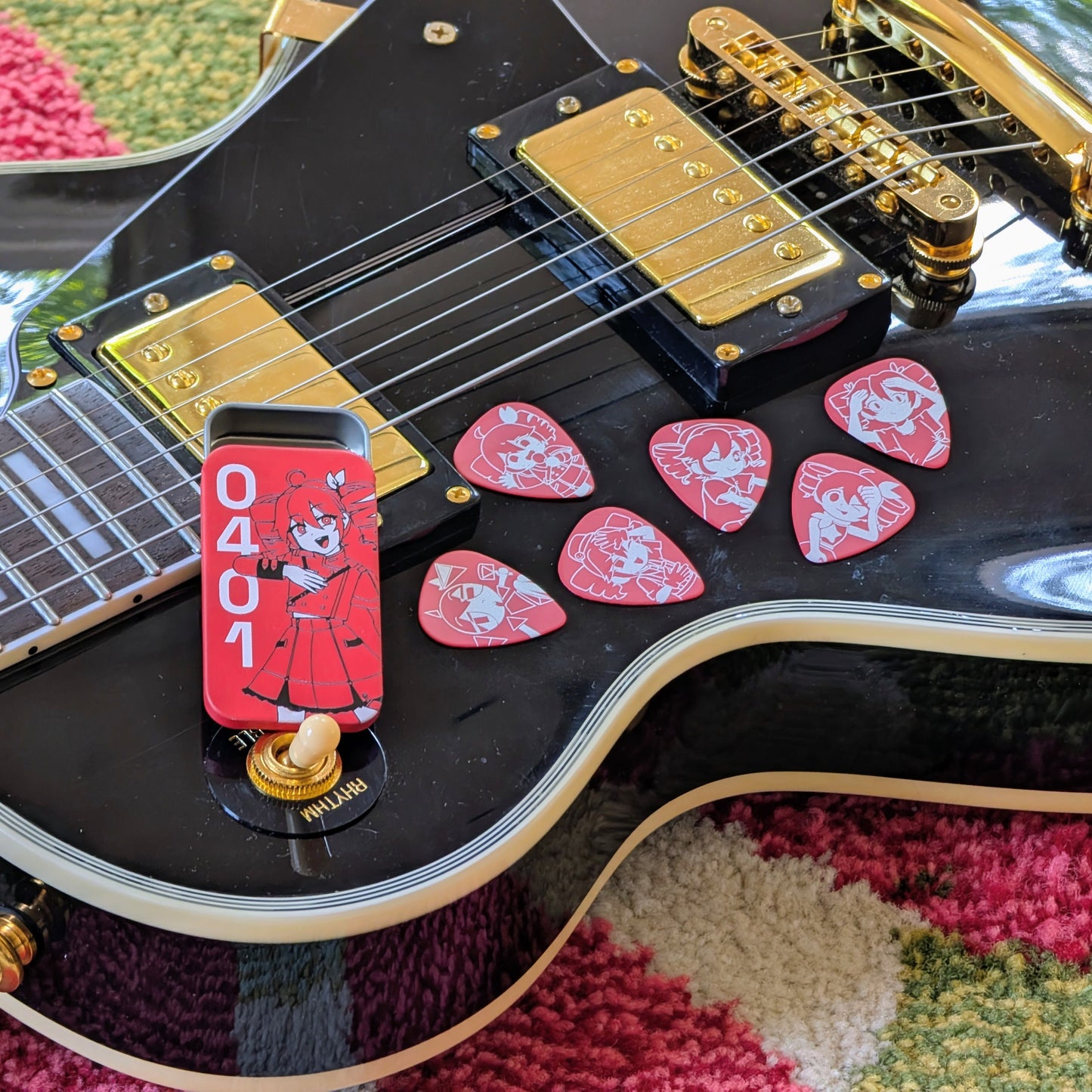 Alternate photo of the first listing image.

A close-up of a black electric guitar body with gold hardware, with several red guitar picks featuring illustrations of Kasane Teto as she appears in different songs and a red metal tin illustrated with a 3/4 Kasane Teto and the numbers "0401".