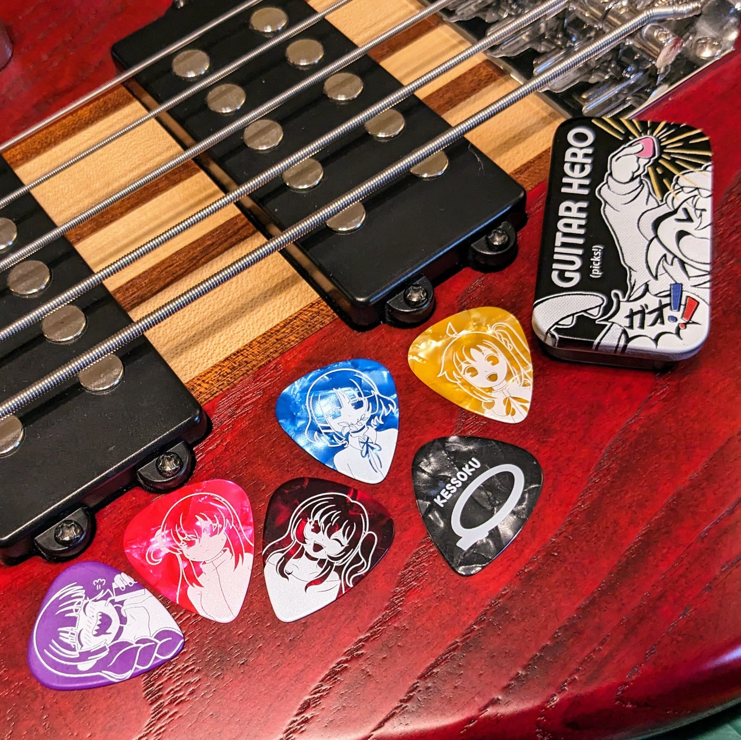 Kessoku Band Guitar Pick set with tin, plus Hiroi Kikuri Bass Pick over a red 5-string bass guitar.